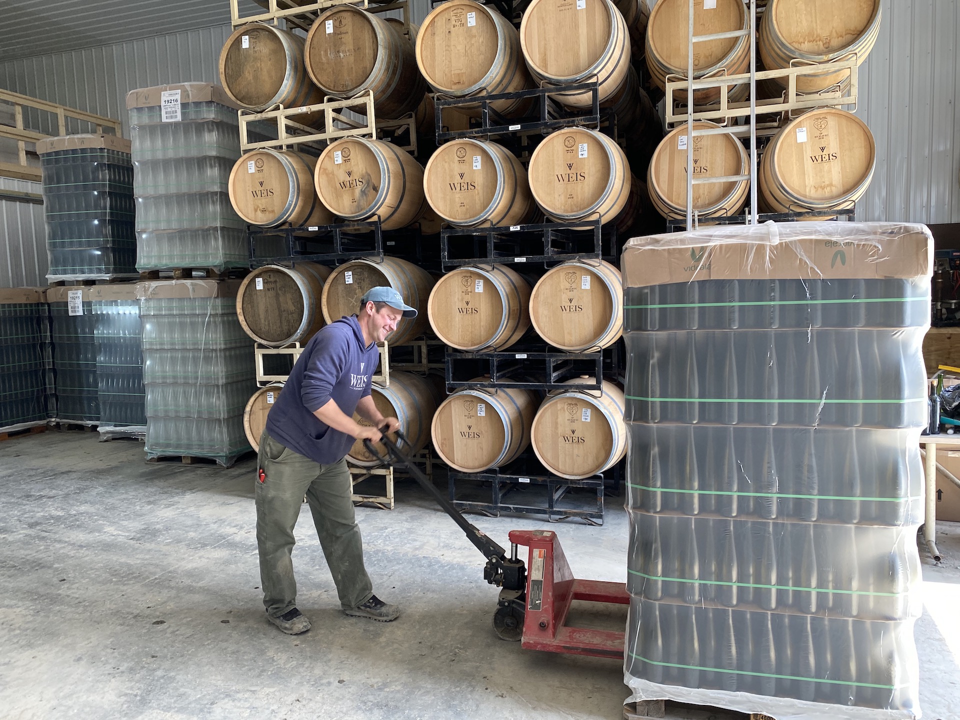 Peter Weis using a pallet jack to move pallets of glass in production facility.  Behind him are stacked wine barrels. 