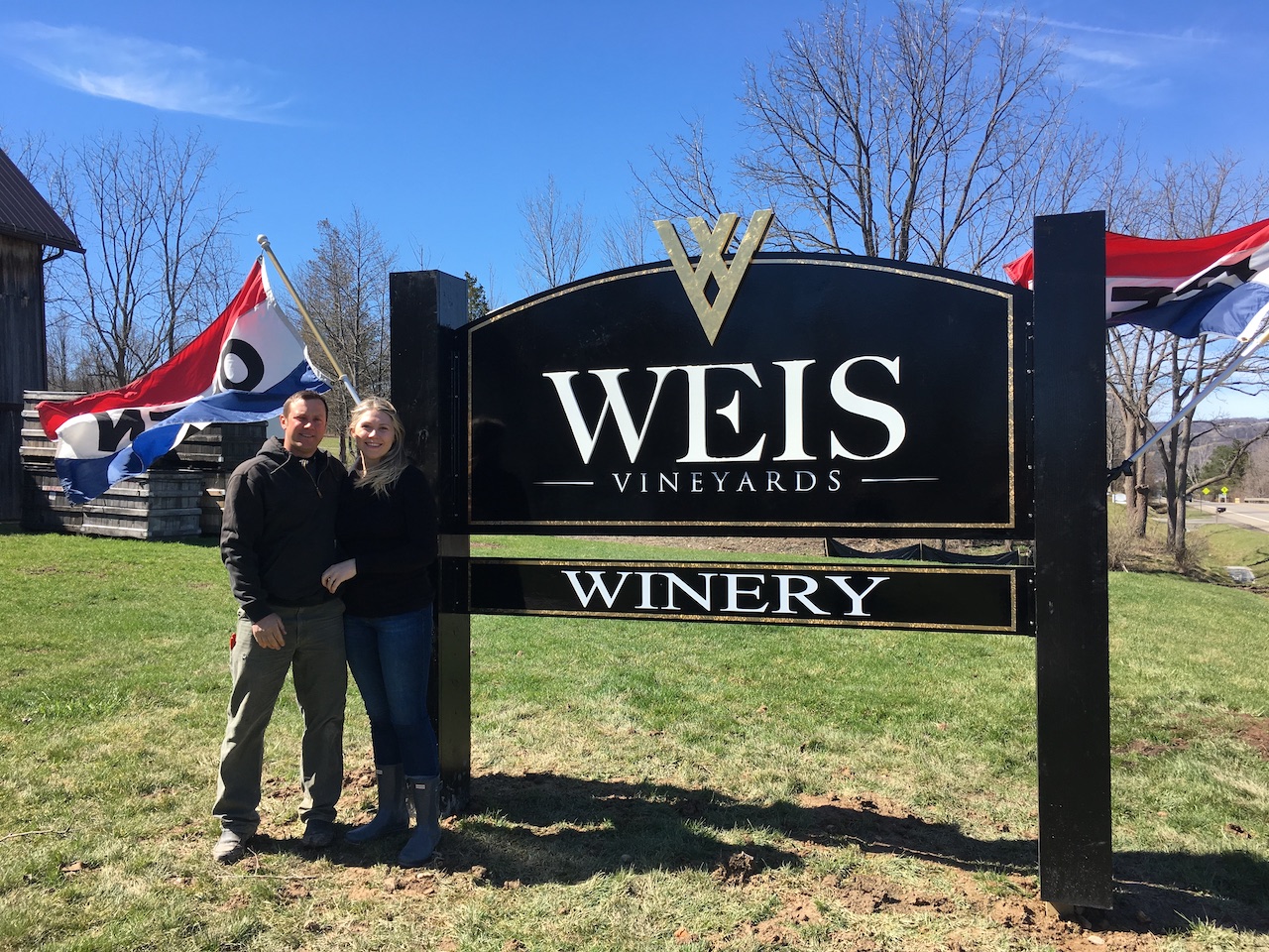 Ashlee and Peter Weis stand in front of the first Weis Vineyards sign in the front yard.  The wind is blowing and two open flags are waving in the background